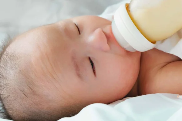 Portrait of a newborn Asian baby on the bed drinks milk from a bottle, Charming black-eyed baby 4 month old lies in bed ,A child resting on a bed