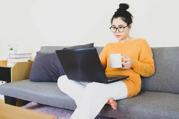 Young beautiful asian woman wore orange knitwear, sitting on gray sofa ,work online lifestyle,relax at home with coffee mug,home working laptop,work from home