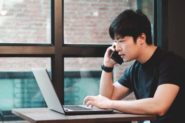 Seçici odak noktası. Zeki Asyalı adam siyah tişört giyiyordu. Laptopla çalışıyor, kahve dükkanında cep telefonuyla konuşuyordu.