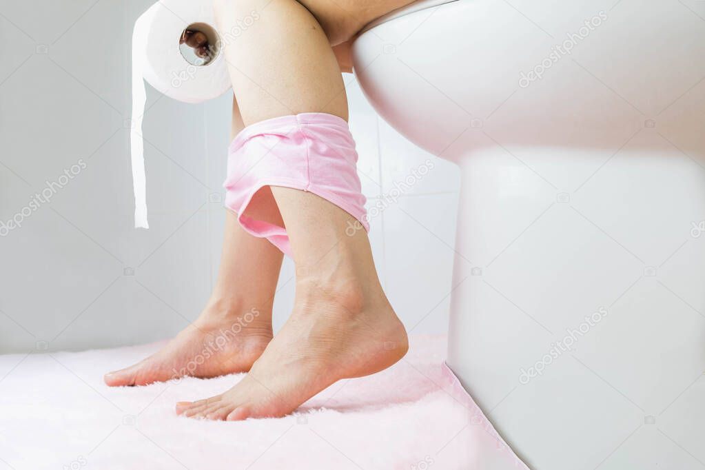 Woman with constipation or diarrhoea sitting on a toilet with her pink panties down around her legs holding unrolled toilet paper ready in her hands, close up view of her legs