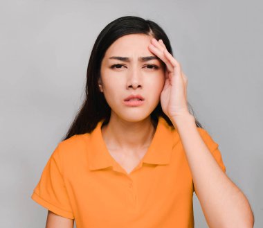 Genç ve güzel Asyalı bir kadın, uzun siyah saçlı, turuncu tişört giyiyor, baş ağrısı ifadesi sergiliyor, gri arka planda kızgın.