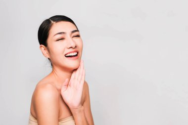 Temiz ve parlak ciltli genç Asyalı kadın, Kozmetik Güzellik Konsepti, yanağına dokunan eller, gri arka planda izole edilmiş gülen ve arkadaş canlısı yüz.