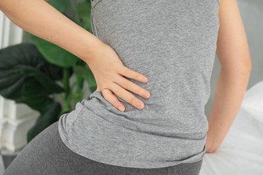 Gri elbiseli genç, güzel Asyalı bir kadın, sabah beyaz bir yatakta uyanır, vücudu ağrır, karın ağrısı, karın ağrısı, ağrı, karın ağrısı ve karın ağrısı ile kıvrılır.