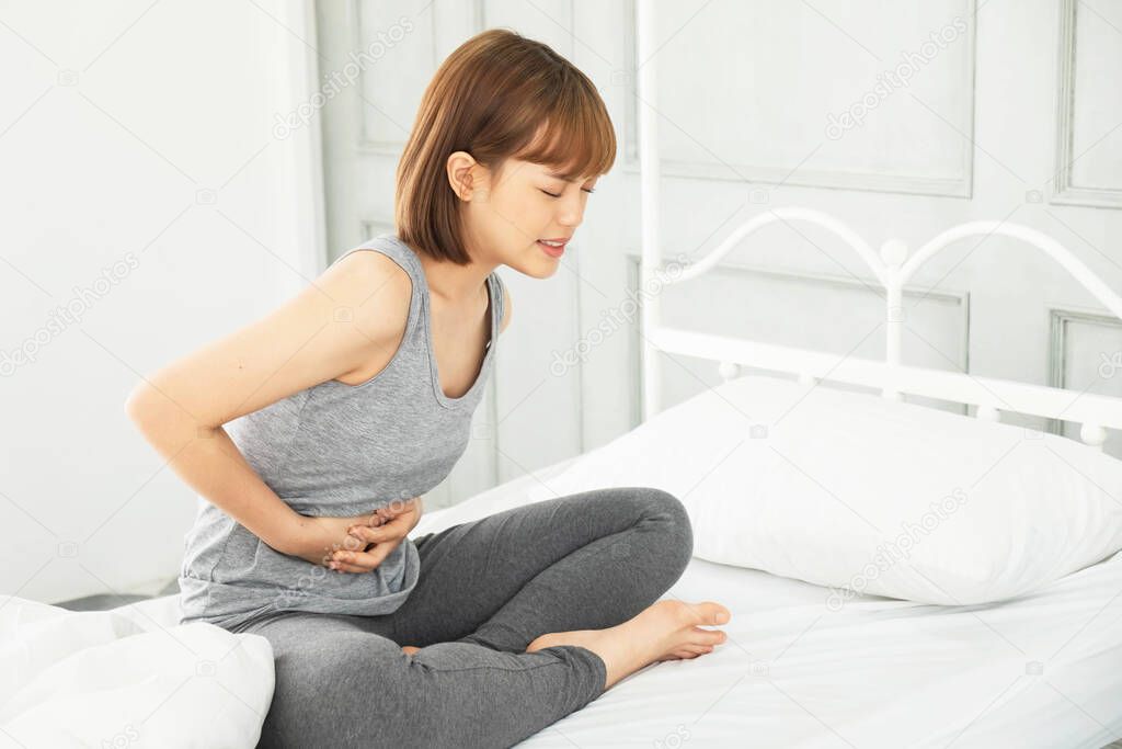 young beautiful asian woman wearing gray clothes,Wake up in a white bed in the morning, The body aches, Hug hands on the stomach Curled up with abdominal pain, stomachache,pain,painful
