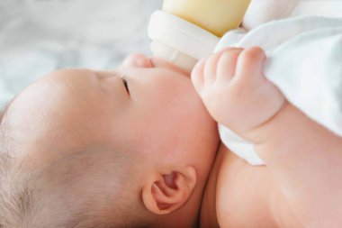 Yatakta yeni doğmuş Asyalı bir bebeğin portresi şişeden süt içiyor, büyüleyici siyah gözlü bebek 4 aylık yatakta yatıyor, bir çocuk yatakta dinleniyor.