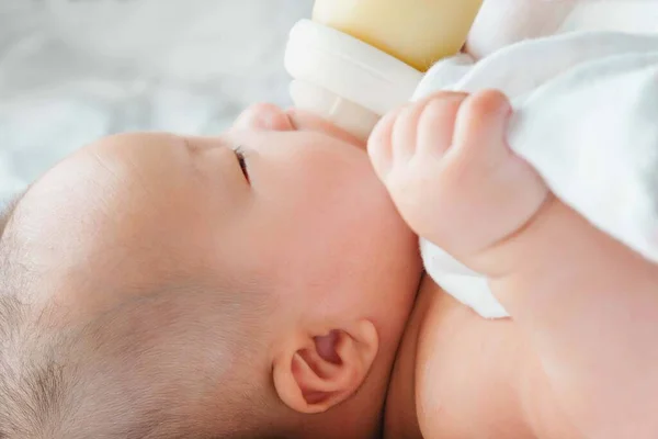 Yatakta yeni doğmuş Asyalı bir bebeğin portresi şişeden süt içiyor, büyüleyici siyah gözlü bebek 4 aylık yatakta yatıyor, bir çocuk yatakta dinleniyor.