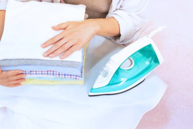 Modern Elektrik Ütüsü ve çamaşır odasındaki pembe arka planda ütü masasında bir yığın kıyafet. Çamaşırları ütüleyen kadın katlanmış ve kurumuş çamaşır yığınları tutuyor.