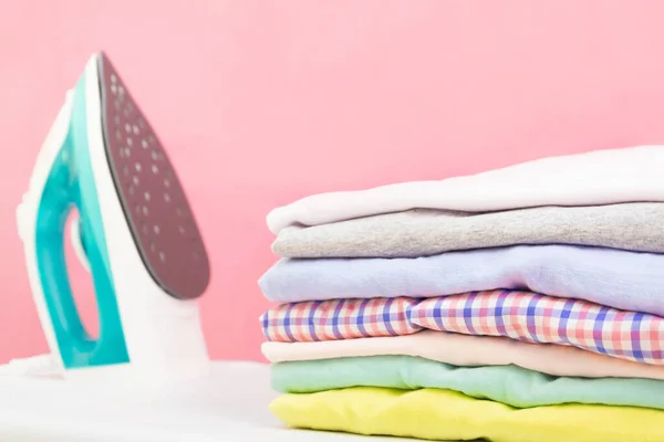 現代の電気アイロンと洗濯室でピンクの背景にアイロン台の服のスタック 家庭用洗濯アイロン洗濯乾燥服の山のスタックを保持女性 — ストック写真
