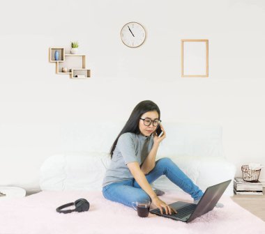 yumuşak seçici odak Genç güzel Asyalı kadın gri t-shirt giydi ve kot pantolon, oturma odasında pembe halı üzerinde oturuyor, online yaşam tarzında çalışıyor, kahve fincanıyla evde dinleniyor, ev bilgisayarı ile çalışıyor, evden çalışıyor.