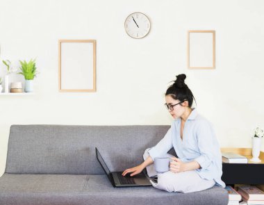Genç, güzel Asyalı bir kadın mavi gömlek giyer, gri koltukta oturur, online yaşam tarzında çalışır, kahve fincanı ile evde dinlenir, evde çalışır, bilgisayarla çalışır, evden çalışır.