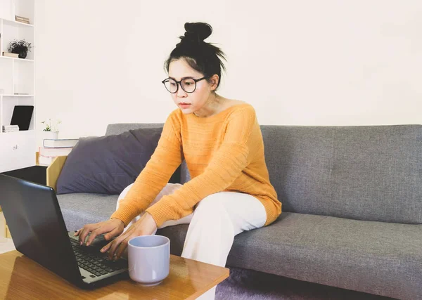 Young beautiful asian woman wore orange knitwear, sitting on gray sofa ,work online lifestyle,relax at home with coffee mug,home working laptop,work from home