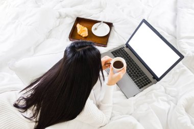 Dizüstü bilgisayarı olan seçici odak kadın, elinde kahve, yatakta yatıyor. en üstteki görünüm