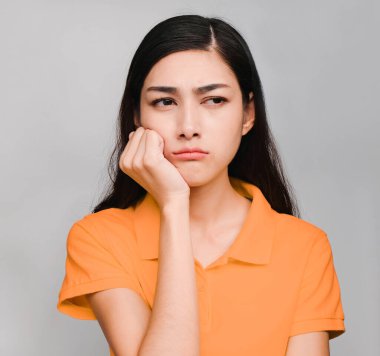Genç, güzel Asyalı kadın, uzun siyah saçlı, turuncu tişört giyiyor, tatmin olmamış bir ifade sergiliyor, gri arka planda yaralanmış.