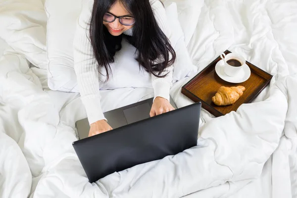 Selective focus woman with a laptop,coffee and Croissant , lying on the bed using and typing on laptop . Girl blogger is typing for a fresh post