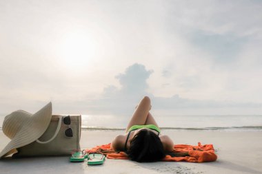 yumuşak seçici odaklı Asyalı kadın kumsalda uzanıyor ve bacaklarını örülmüş çantalar, şapka ve güneş gözlüklerinin yanındaki gökyüzü günbatımının, yaz tatilinin ve tatil konseptinin fotokopi alanına doğru çeviriyor.