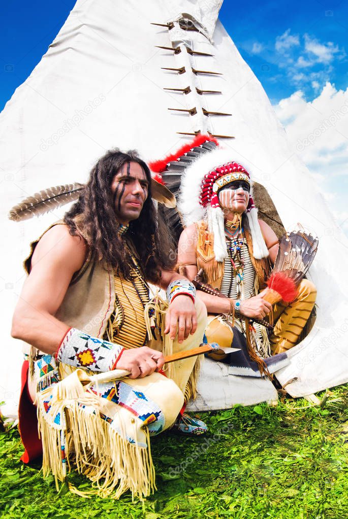 Two american indian sits beside wigwam