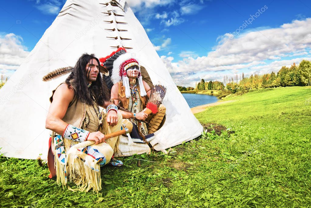 Two american indian sits beside wigwam