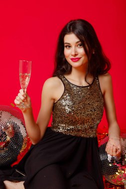 Makyajlı ve kırmızı dudaklı mutlu kız yerde oturuyor. Siyah elbiseli, altın pullu ve şampanyalı, kırmızı arka planda altın konfetileri olan disko toplarıyla.