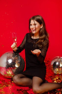 Makyajlı ve kırmızı dudaklı mutlu kız siyah elbiseli disko toplarıyla yerde oturuyor ve şampanya içiyor, kırmızı arka planda altın konfetileri uçuşuyor.
