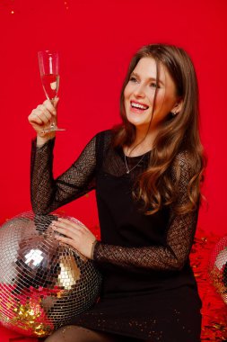 Makyajlı ve kırmızı dudaklı mutlu kız siyah elbiseli disko toplarıyla yerde oturuyor ve şampanya içiyor, kırmızı arka planda altın konfetileri uçuşuyor.