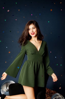 Kırmızı dudaklı yeşil elbiseli güzel esmer bir kızın portresi konfeti yağmurunda duruyor ve mavi bir arka planda kutlama yapıyor.