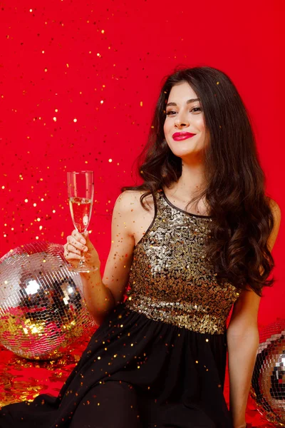 Happy girl with make-up and red lips sits on the floor with disco balls in a black dress with gold sequins and drinks champagne, gold confetti fly on a red background