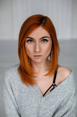 Kırmızı saçlı, gri kazaklı, omuzları açık genç bir kadın kameraya bakıyor.