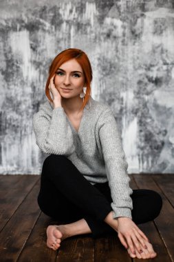 Gri tişörtlü, omuzları açık kızıl saçlı genç bir kadın yerde oturmuş kameraya bakıyor.