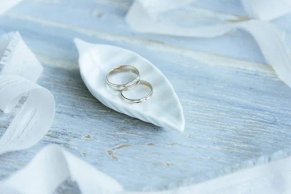 Dos anillos de boda de oro amarillo y blanco en una hoja blanca decorativa de cerámica y una mesa de madera azul, cinta vintage decorativa blanca alrededor, accesorios para novias, estilo boho rústico floral . —  Fotos de Stock