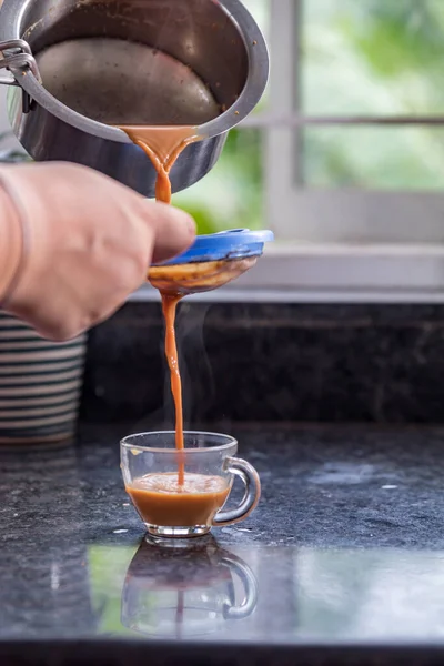 Plastik süzgeçten geçip cam fincana buharlı masala çayı döken bir el.
