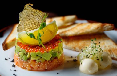 Kırmızı havyar, avokado ve balıklı tartar. Tartar beyaz bir tabakta duruyor ve yanında da kızarmış ekmekle tereyağı var. Balık pulu ve limonla süslenmiş..