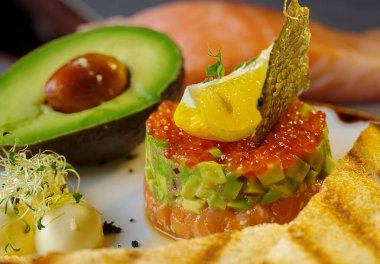 Kırmızı havyar, avokado ve balıklı tartar. Tartar beyaz bir tabakta duruyor ve yanında da kızarmış ekmekle tereyağı var. Balık pulu ve limonla süslenmiş..