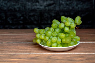 Tahta üzerinde koyu arkaplan olan beyaz bir tabakta çekirdeksiz üzüm.