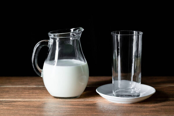 An empty glass cup and a jar with cow's milk on a wooden table and dark background