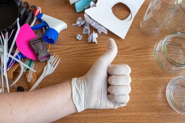 Person shows thumbs up by hand in latex glove. On the wooden table lies trash sorted into three groups: plastic, paper and glass. Waste sorting theme.