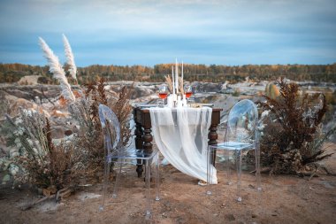 Düğün süslemeleri. Açık hava düğün yeri kurumuş çiçek kompozisyonu, şarap ve mumlarla dolu bir gala masası ile süslenmiştir. Masanın yanında şeffaf plastikten yapılmış iki sandalye var.