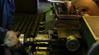 Fabrikada, eldivenli bir işçi metal işlerinin işlendiği torna tezgahını kontrol eder. Metal parçalar işyerinden kesilmiş. İş parçası klemplenir ve büyük bir varile konur ve sıcak bir ışıkla aydınlatılır. Yan görünüm. 