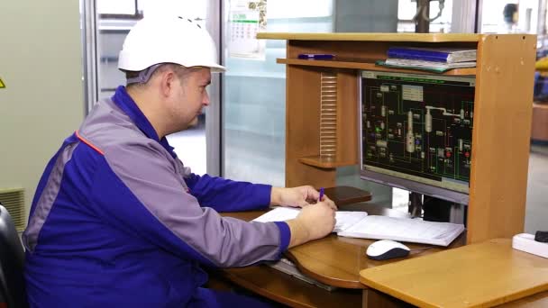 实验室里穿着蓝色制服 头上戴着白色头盔的专家坐在桌旁 在一本书中写作 桌上有一台电脑 上面有工作设备的图表 — 图库视频影像