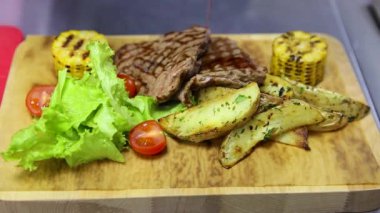 Şef yemeği ahşap bir tabakta kırmızı sosla suluyor. Tabakta salata yaprakları, kızarmış patates dilimleri, et ve domates ve mısır koçanı yer alıyor. Yakın plan.