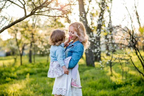 Happy mom and daughter are walking on the street in the garden, in the park, hugging. Mom kisses her daughter. Daughter kisses mom. Green grass, flowering trees in the garden. Mothers Day