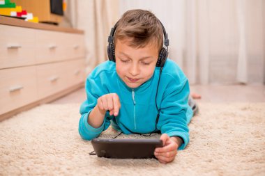 Çocuk evde dizüstü bilgisayar ve tabletle yerde yatıyor. Çevrimiçi öğrenme. Karantina ve tecrit. Reyonok başka bir online tasarımcıyla oynuyor. Evde eğitim. Kulaklıklı bebek gülümsüyor.