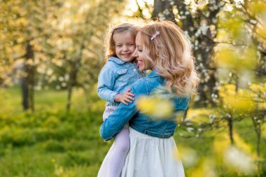 Mutlu, sarışın anne ve kız bahçede, parkta, kucaklaşarak yürüyorlar. Anne kızını öpüyor. Kızı anneyi öpüyor. Yeşil çimenler, çiçek açan ağaçlar. Anneler Günü. Kızların günü..