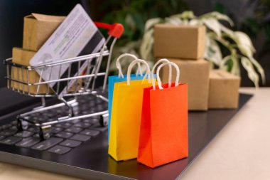 İnternet alışveriş konsepti. Küçük alışveriş arabası, kutular, renkli çantalar, banka kartı, dolarlar dizüstü bilgisayarda duruyor.