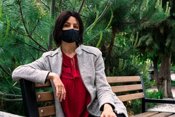 Coronavirus modaya uygun bir tıbbi yüz maskesi. Parktaki bankta oturan genç siyah saçlı kızlar tarafından takılıyor. Gri bir ceket ve kırmızı bluz giyiyorlar.
