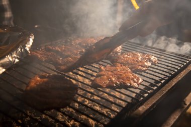 Akşamları lezzetli bifteklerin açık ateşte kızardığı ızgarada bir el maşayı tutar ve eti çevirir. Yakın çekim, yumuşak odaklanma. duman altı çizili