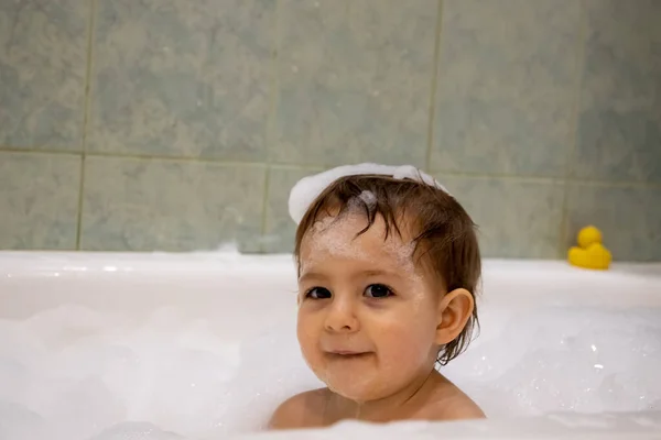Мила маленька дівчинка посміхається, дивлячись на камеру, приймаючи ванну з мильною піною під кутом і головою. крупним планом, м'який фокус, фон зелена ванна кімната в розмитому . — стокове фото