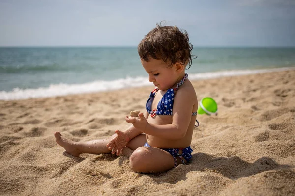 Mała słodka dziewczynka maluch w niebieskim stroju kąpielowym na piaszczystej plaży bawi się w piasek na tle fal morskich w ciepły słoneczny dzień. — Zdjęcie stockowe