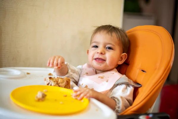 Lindo bebé se sienta en una silla de bebé naranja con una mesa con las manos y la cara sucias, se ve en la cámara y se ríe. sobre la mesa hay migajas y un plato de color naranja. primer plano, vista lateral, enfoque suave, fondo borroso — Foto de Stock