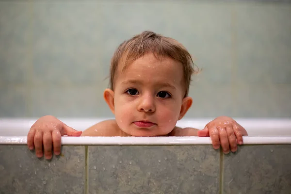 Cute Caucasian baby peeks out of the bathtub, put hands on the side of the bath and looks to the camera tired. in the background is a green bathroom in blur. close-up, soft focus Stock Photo