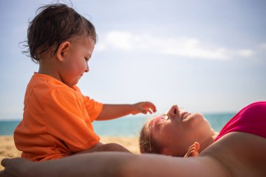 Küçük tatlı çocuk kumsalda yatan annesini rahatsız ediyor ve oyun oynuyor. Arka planda güneşli bir günde ılık deniz